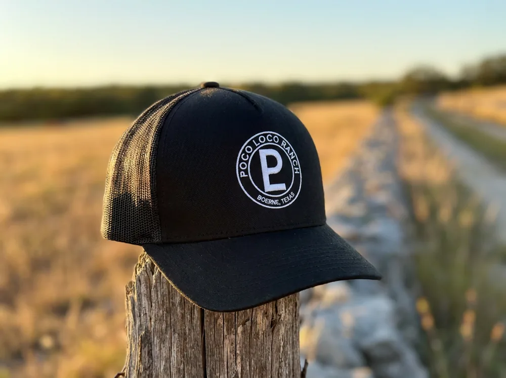 Poco Loco Ranch trucker hat in black, on a fence post along the ranch road